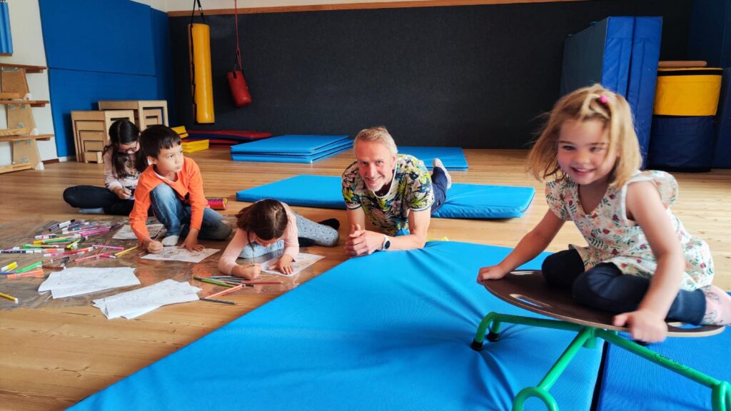 Kinder beim Kindergottesdienst Schwaz: Eine fröhliche Gruppe von Kindern und einem Erwachsenen beim gemeinsamen Malen und Spielen auf Turnmatten in einem hellen Raum.